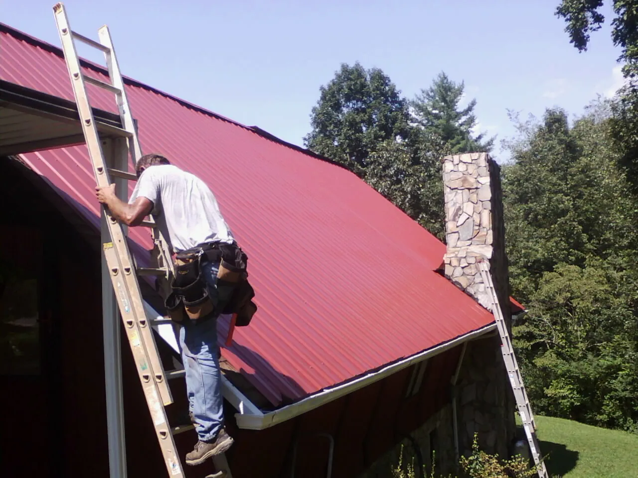 Roofer on a ladder beside a red metal roof and stone chimney — Soffit & Fascia Repair by Roofing Brothers in Esmond